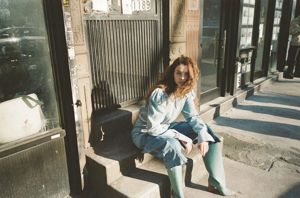 A young woman with long, wavy red hair sits on stone steps outside a building, wearing a light blue denim outfit and green boots in sunlight.