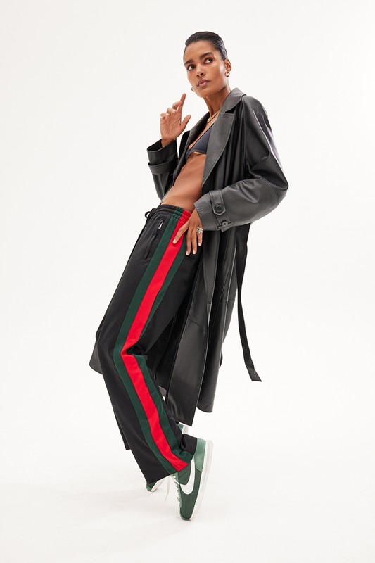 Fashion photo of a woman posing in a black coat, bra top, track pants with red and green stripes, and green sneakers against a white backdrop.