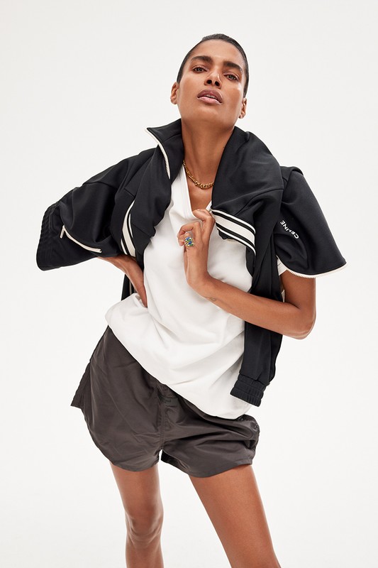 An adult female model in a white shirt, black jacket, and gray shorts poses against a white background in a fashion shot.