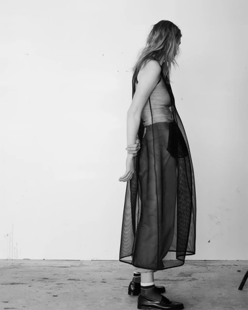 Monochrome portrait of a slender woman in a sheer vest and skirt, posing against a plain white wall in black leather shoes and socks.