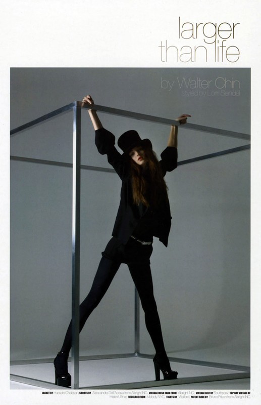 Editorial fashion shot of a woman in a black top hat and dark clothing posing in a large metal cube in a light grey studio.