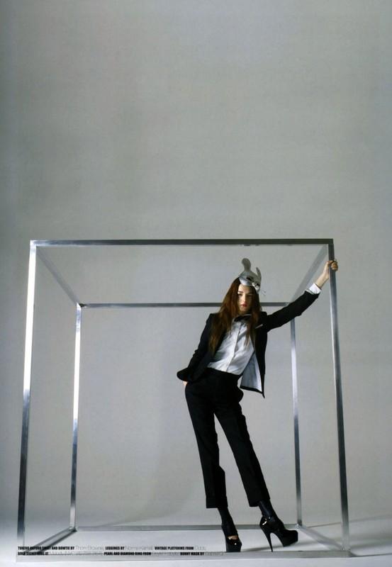 A woman in a black suit and heels poses inside a silver cube with a futuristic headpiece in a studio, creating a stylish and surreal image.