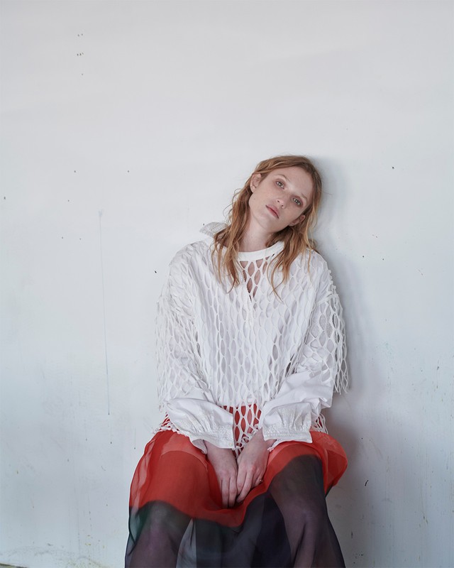 An adult female model is sitting in a white textured top with long sleeves and a long red skirt against a white concrete wall.
