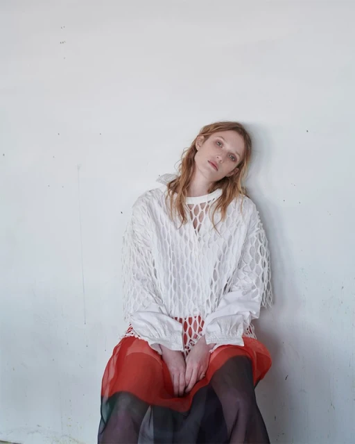 An adult female model is sitting in a white textured top with long sleeves and a long red skirt against a white concrete wall.