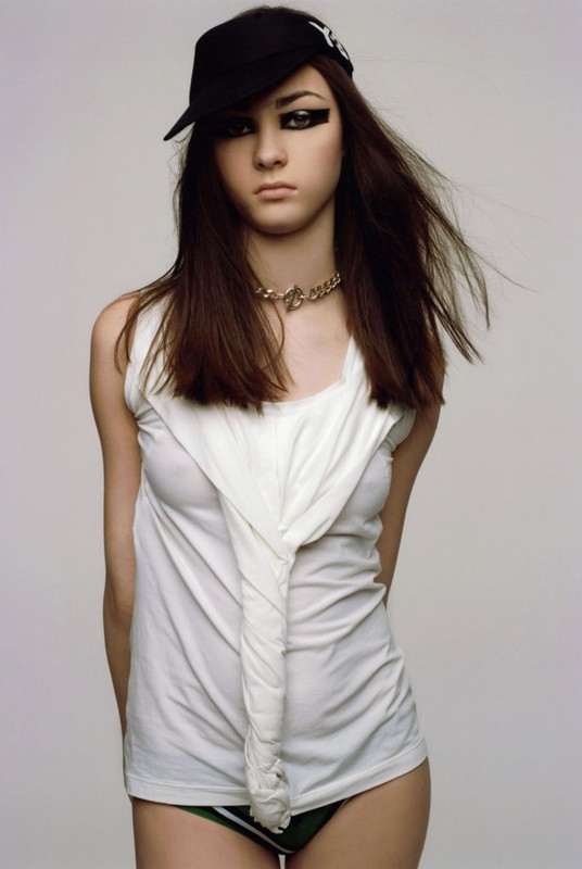 Teen girl with long brown hair poses in a white tank top and black baseball cap against a light gray backdrop in a studio shot.