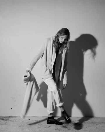 Model wearing a knit cardigan and trousers holding an extra sweater against a wall with strong shadows