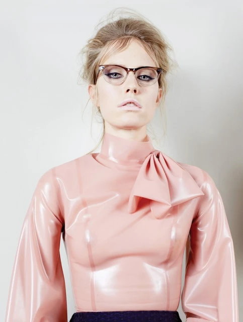 Blond woman in a glossy pink blouse with bow and glasses posing against a neutral background in a studio portrait.