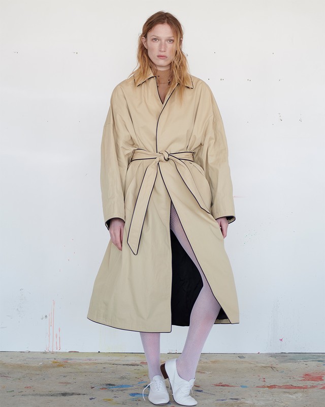 A full shot of a redhead adult female standing in a long beige trench coat with black piping and white tights against a white wall.