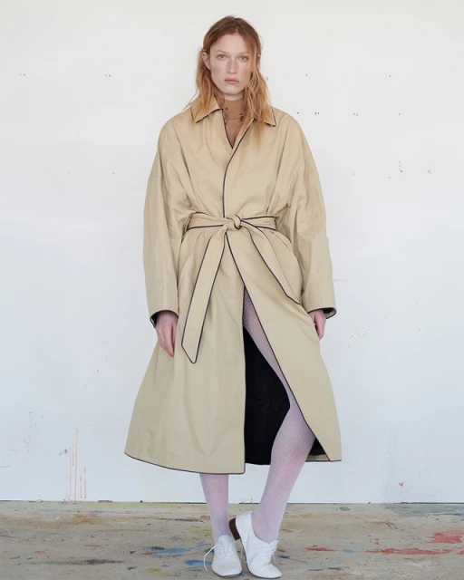 A full shot of a redhead adult female standing in a long beige trench coat with black piping and white tights against a white wall.