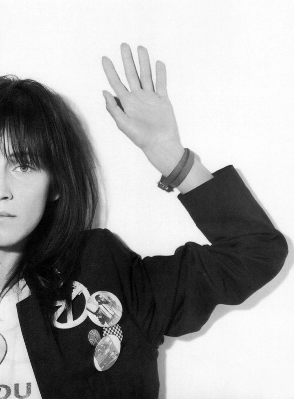 Monochrome portrait of a young adult woman with dark hair, wearing a blazer with buttons and raising her hand against a plain white wall.