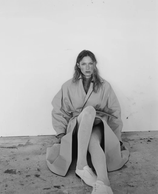 A woman with long hair sits indoors wearing a tan trench coat, white shoes, and textured stockings against a white wall.
