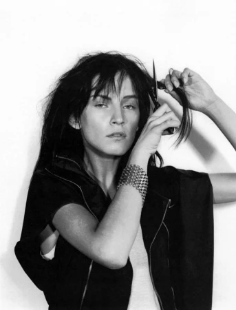 Monochrome portrait of a woman cutting her dark hair with scissors, wearing a dark jacket and a wide metallic cuff bracelet.