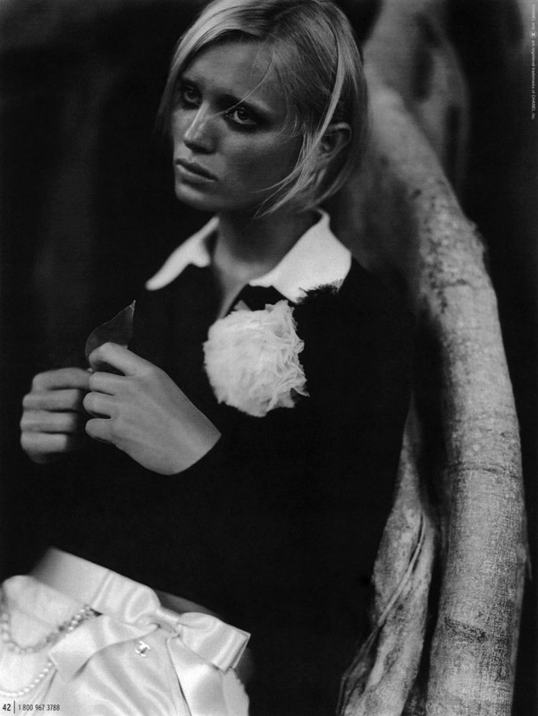Monochrome portrait of a fair-skinned young woman with short blonde hair, wearing a dark shirt and white satin shorts.