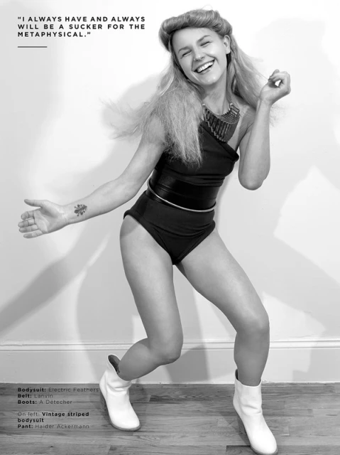 Black and white photo of a young woman in a bodysuit and boots, laughing and dancing in front of a plain wall in a studio setting.