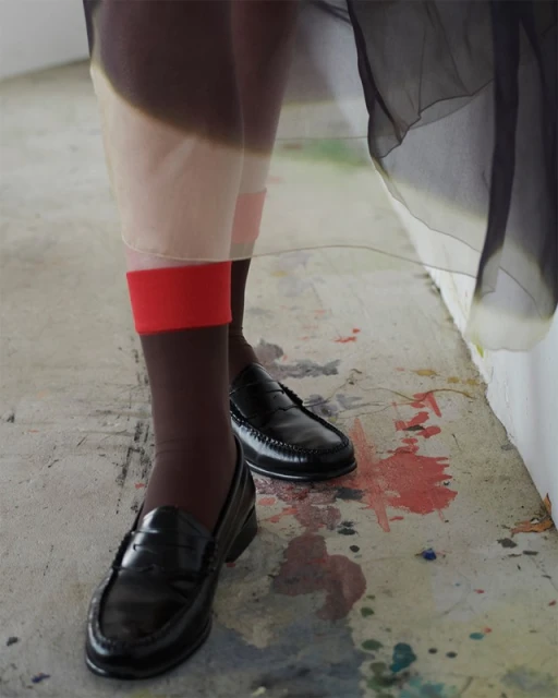 Low-angle shot of a person in sheer skirt, red socks, and black loafers stands on a concrete floor with paint splatters. Fashion editorial.