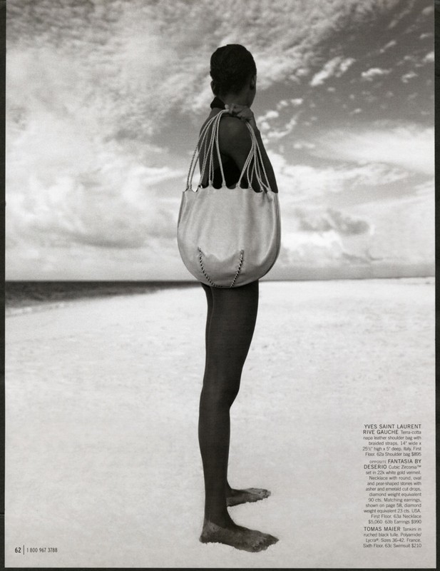 Fashion shot of a woman standing on a sandy beach with a large tote bag, against a background of sky and water.