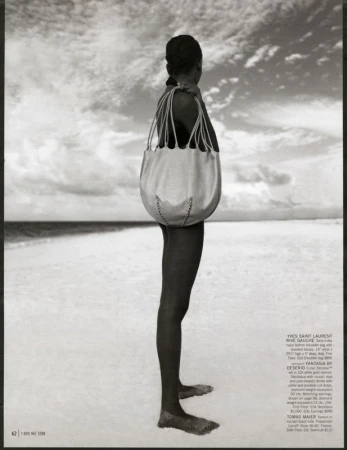 Fashion shot of a woman standing on a sandy beach with a large tote bag, against a background of sky and water.