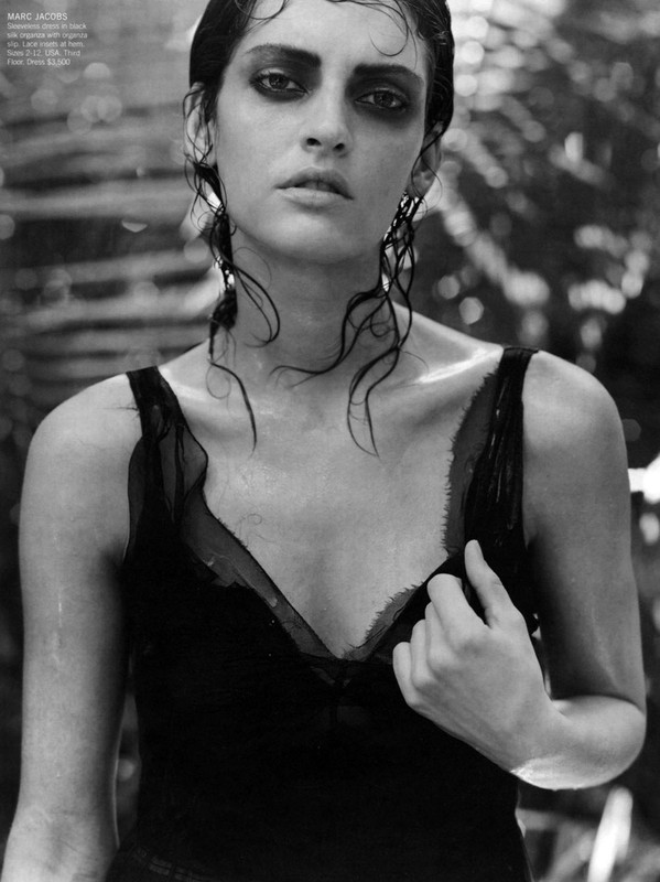Monochrome portrait of a woman with dark eye makeup wearing a black top, wet hair, and dangling earrings, set against a blurred background.