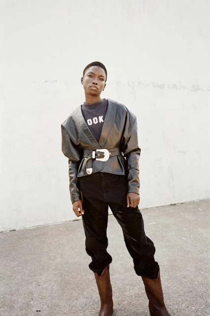 Fashionable portrait of an adult woman wearing a belted black leather jacket, pants, and brown boots standing against a wall.