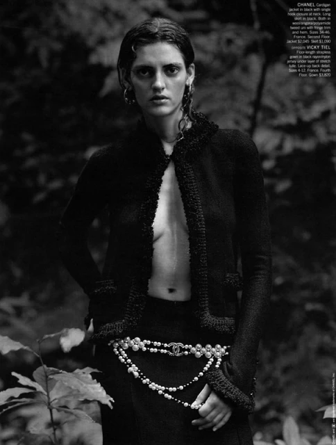 Monochromatic outdoor fashion shot: woman in black jacket and pearl belt poses in forest. Moody lighting and soft focus.