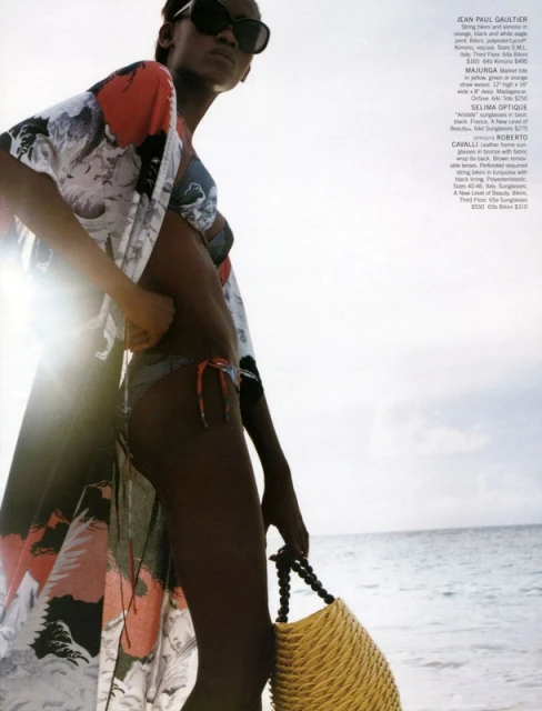 Model in bikini and floral cover-up poses on a beach with a straw bag, wearing sunglasses, against a sky and ocean backdrop.