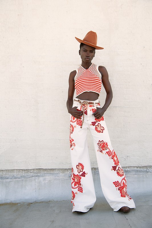 An adult woman poses in a red and white striped crochet crop top and white jeans with red floral detail in a fashion editorial shot.