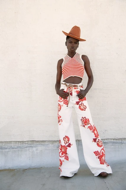 An adult woman poses in a red and white striped crochet crop top and white jeans with red floral detail in a fashion editorial shot.