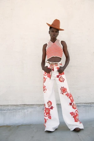 An adult woman poses in a red and white striped crochet crop top and white jeans with red floral detail in a fashion editorial shot.