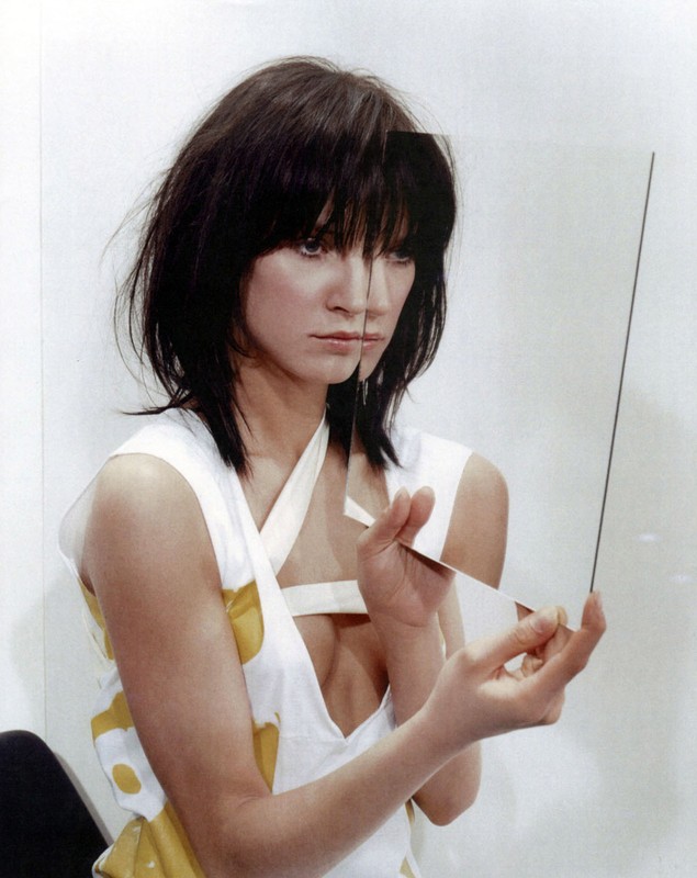 Brunette woman with unique hairstyle holds glass panes in a photo studio, wearing a white top with yellow accents in a portrait.