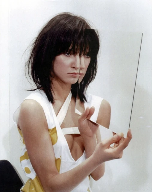 Brunette woman with unique hairstyle holds glass panes in a photo studio, wearing a white top with yellow accents in a portrait.
