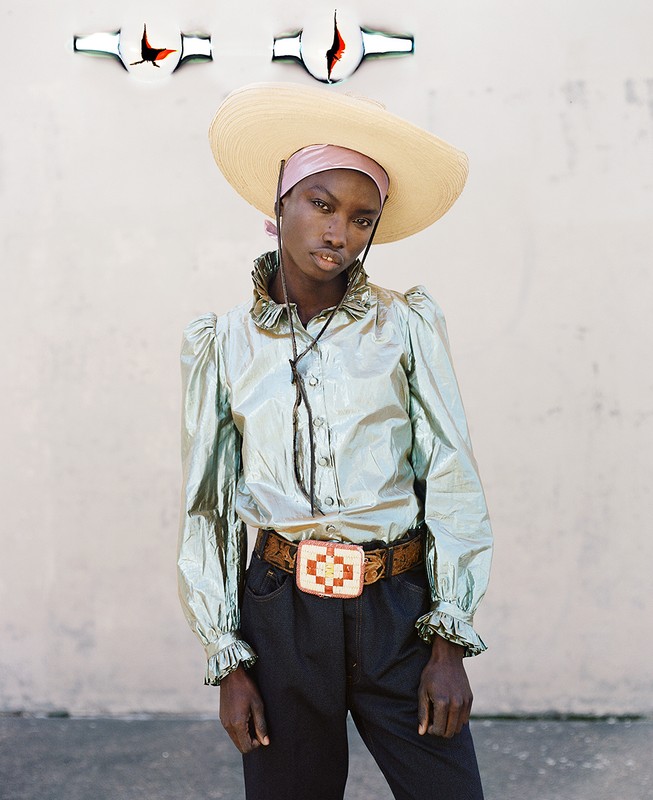 Fashion shot of a Black woman wearing a large hat, a shiny collared shirt, and a belt with a large buckle against a neutral wall.