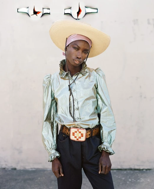 Fashion shot of a Black woman wearing a large hat, a shiny collared shirt, and a belt with a large buckle against a neutral wall.