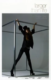 Model wearing black top hat and dark outfit holding onto a large metallic cube frame in a studio setting.