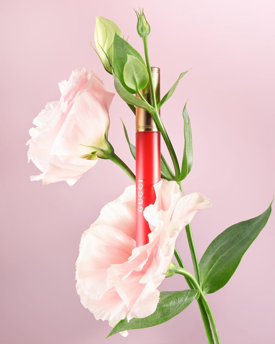 A vibrant red lipstick tube nestled amongst blooming light pink flowers and green stems against a solid pale pink background.