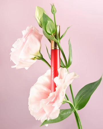 A vibrant red lipstick tube nestled amongst blooming light pink flowers and green stems against a solid pale pink background.