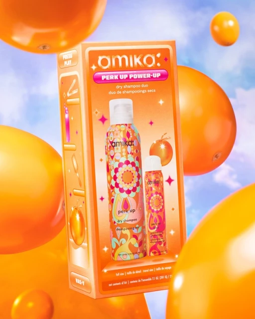 Amika Perk Up Power-Up dry shampoo duo in orange packaging with floating orange orbs against a blue sky, for haircare product shot.