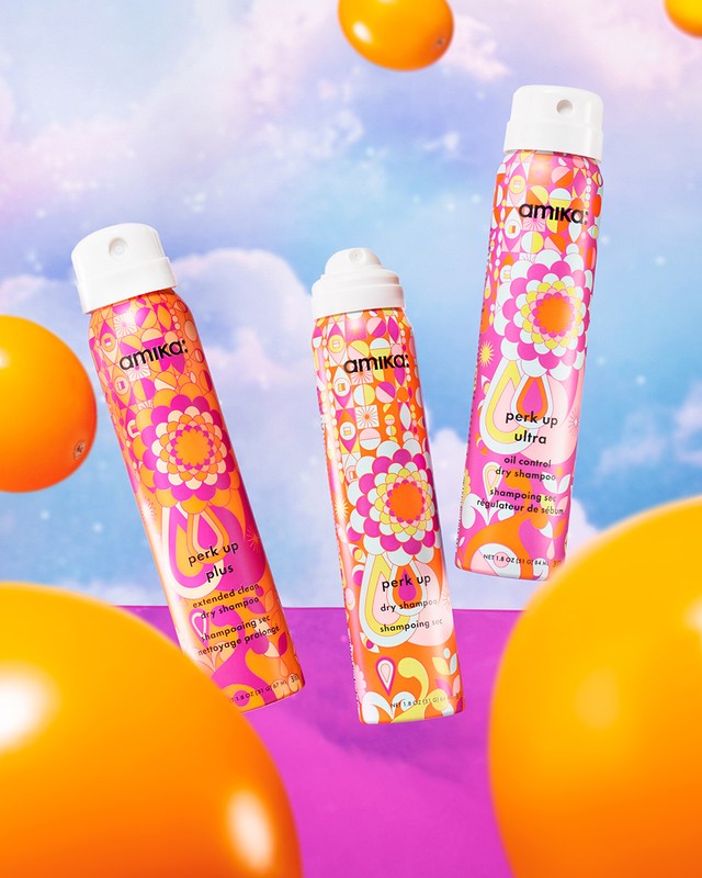 Three Amika dry shampoo bottles with orange geometric floral pattern on a violet background with bright orange round objects.
