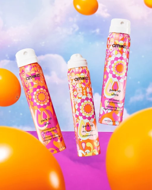 Three Amika dry shampoo bottles with orange geometric floral pattern on a violet background with bright orange round objects.