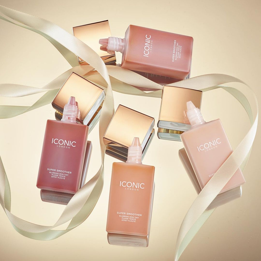 Several bottles of Iconic London Super Smoother with gold lids, arranged on a reflective surface with ribbon accents for beauty product promotion.