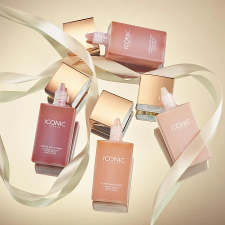Several bottles of Iconic London Super Smoother with gold lids, arranged on a reflective surface with ribbon accents for beauty product promotion.
