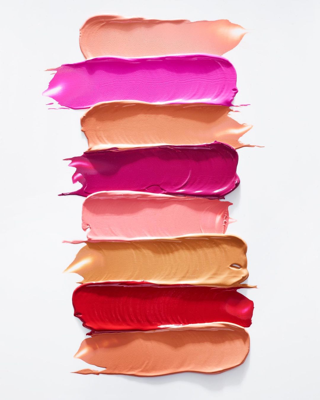 Close-up of colorful lipstick swatches on a white surface, showcasing a variety of pink, red, and peach tones in thick, glossy strokes.
