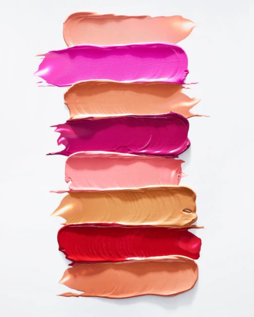 Close-up of colorful lipstick swatches on a white surface, showcasing a variety of pink, red, and peach tones in thick, glossy strokes.