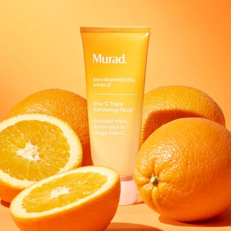 Murad Vita-C Triple Exfoliating Facial tube surrounded by whole and halved oranges on an orange backdrop, skincare product shot.