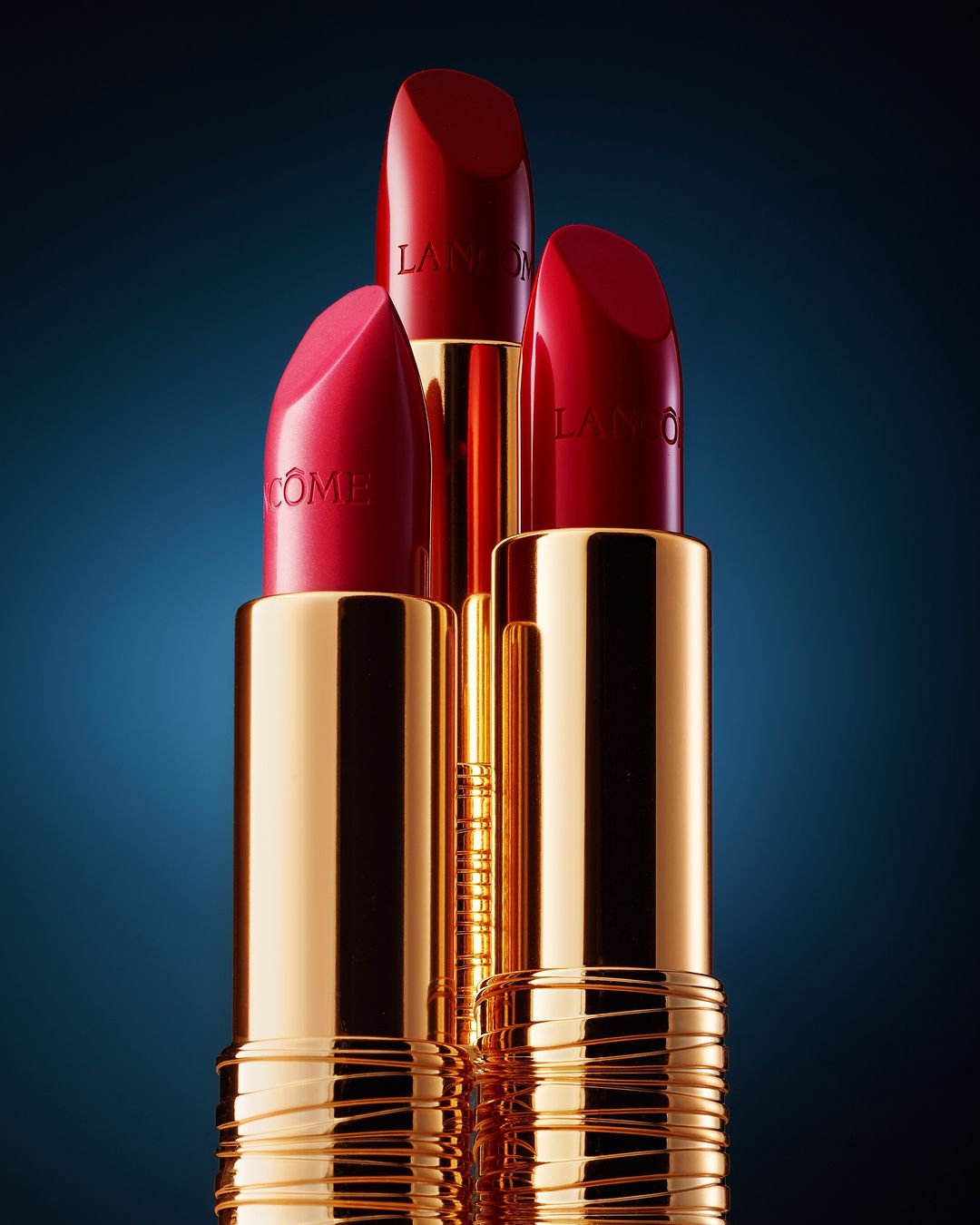 Three red lipsticks stand prominently, showcasing gold casings and the Lancôme logo against a dark blue gradient background. Close-up beauty product.