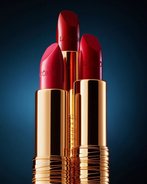 Three red lipsticks stand prominently, showcasing gold casings and the Lancôme logo against a dark blue gradient background. Close-up beauty product.