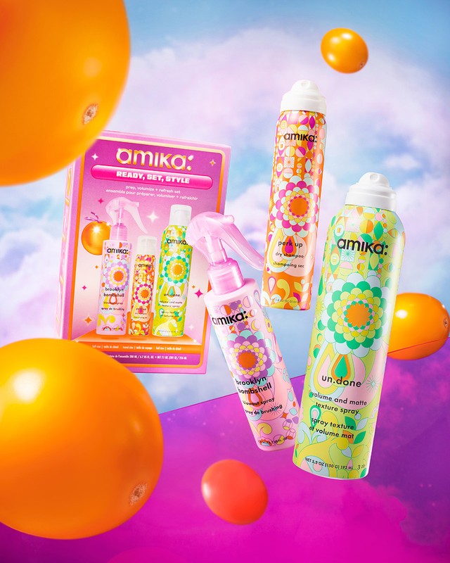 Amika hair product gift set with colorful packaging and floral patterns, set against a blurred sky background with floating yellow fruit.