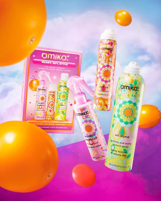 Amika hair product gift set with colorful packaging and floral patterns, set against a blurred sky background with floating yellow fruit.