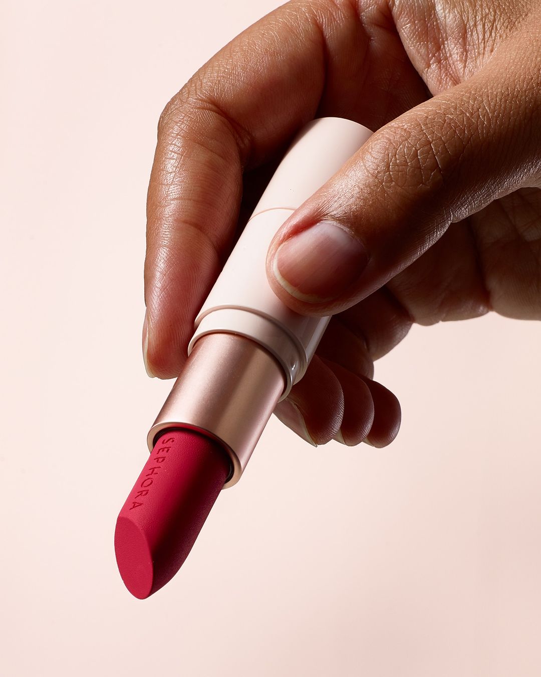 A close-up shot of an adult human hand holding a pink lipstick with the SEPHORA logo, set against a soft pink background. Beauty product.