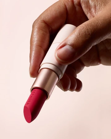 A close-up shot of an adult human hand holding a pink lipstick with the SEPHORA logo, set against a soft pink background. Beauty product.