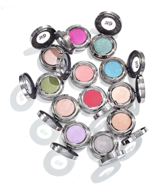 A collection of colorful Urban Decay eyeshadow singles are displayed on a white background, showcased in their sleek, reflective silver compacts.